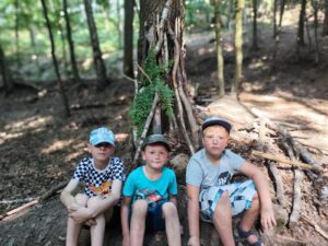 Kluci se fotí v lese u postaveného domečku z větví a listí na dětském letním táboře.