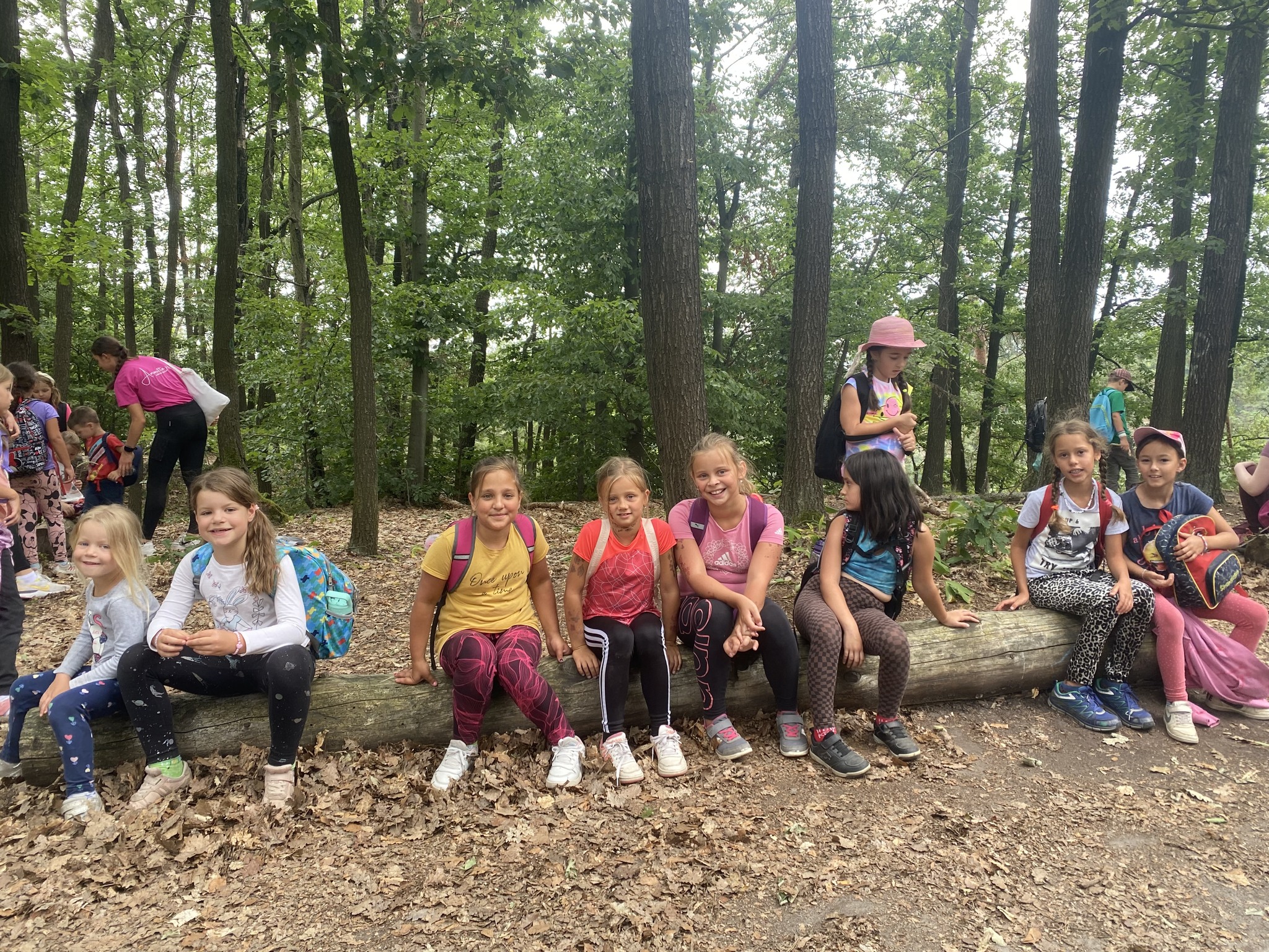 Děti na letním dětském táboře sedí na velkém kmeni v lese a odpočívají během výletu.