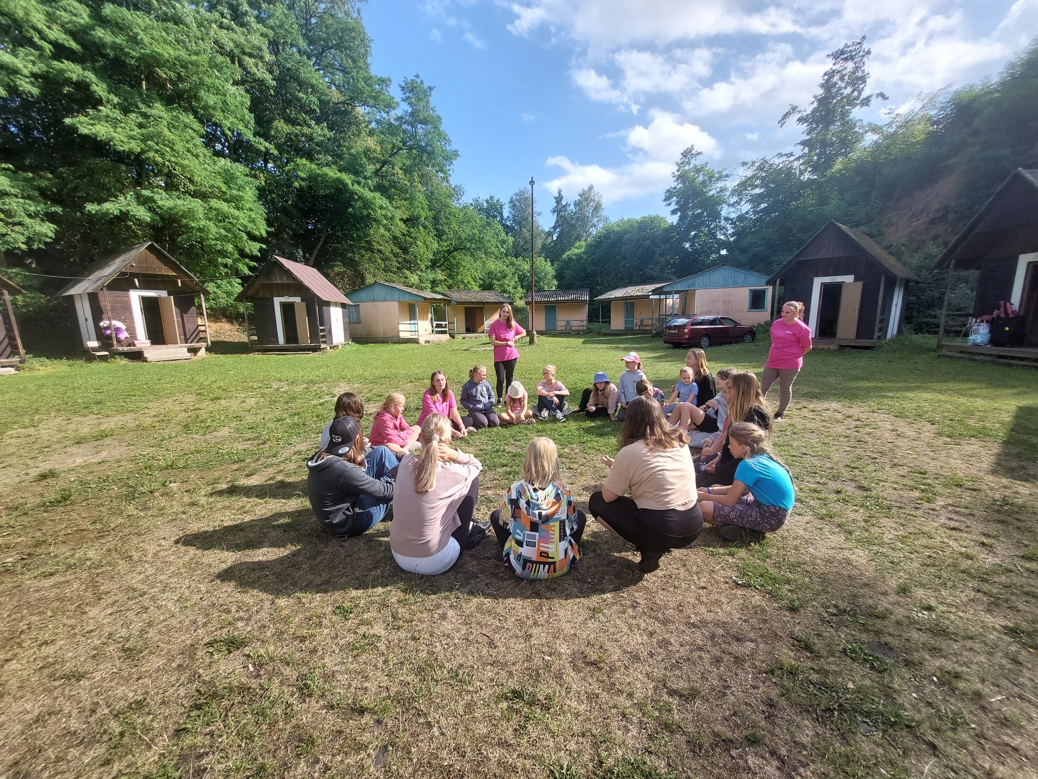 Děti na letním klasickém táboře sedí v kroužku u táborových chatek a povídají si společně.