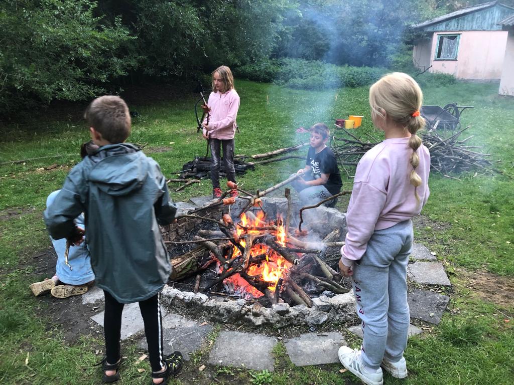 Děti na letním dětském táboře stojí u ohně, opékají špekáčky a dívají se na hvězdy.